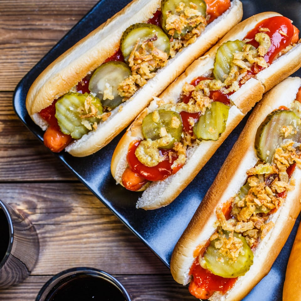 Three hot dogs with ketchup, mustard, pickles, and crispy onions on a black plate.